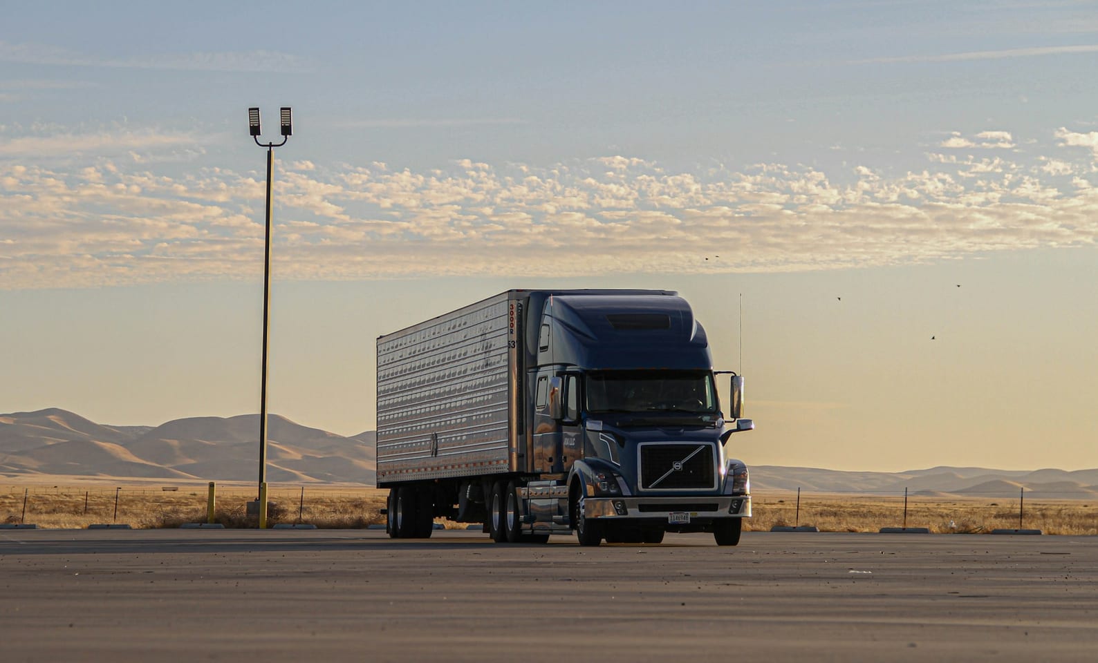 A blue tractor and ribbed trailer parked on an empty lot at first light, low hills and open sky behind